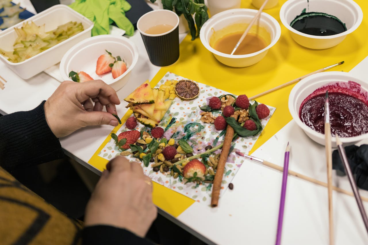 Hands arranging fruits and ingredients creatively during food art preparation indoors.
